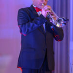 Mężczyzna w ciemnym garniturze gra na trąbce. Tło jest rozmyte, w odcieniach niebieskim i różowym. Jasne oświetlenie podkreśla scenę.