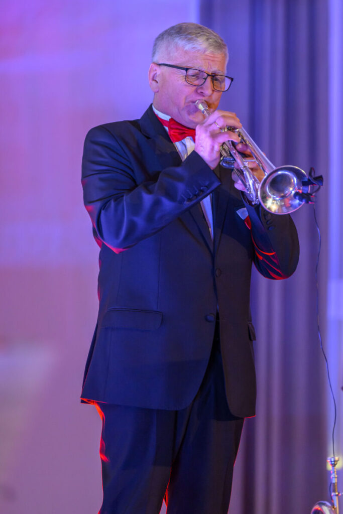 Mężczyzna w ciemnym garniturze gra na trąbce. Tło jest rozmyte, w odcieniach niebieskim i różowym. Jasne oświetlenie podkreśla scenę.