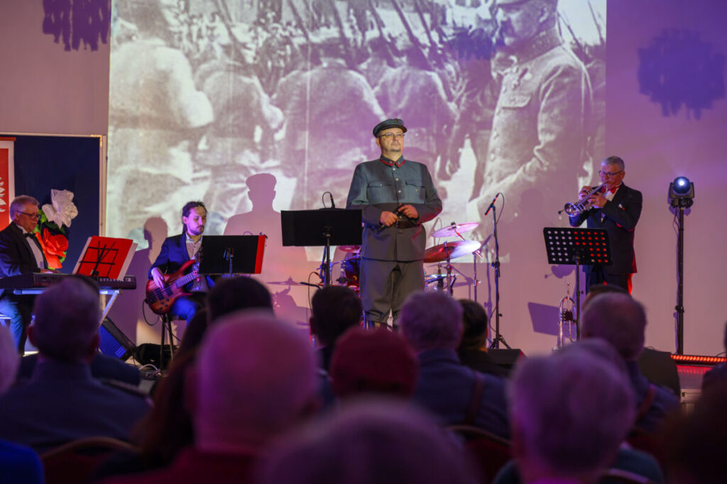 Na zdjęciu widać scenę z grupą muzyków, w tym jedną osobę w wojskowym mundurze. Tło jest kolorowe, z historyczną projekcją. Widownia składa się z kilku osób, niektóre oświetlone punktowym światłem.