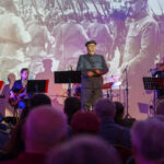 Na zdjęciu widać scenę z grupą muzyków, w tym jedną osobę w wojskowym mundurze. Tło jest kolorowe, z historyczną projekcją. Widownia składa się z kilku osób, niektóre oświetlone punktowym światłem.