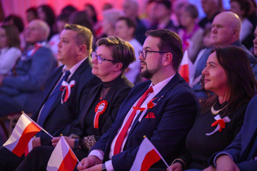 Grupa elegancko ubranych osób siedzi, trzymając polskie flagi. W tle widoczna rozmyta publiczność w odcieniach czerwieni. Wszyscy mają biało-czerwone kotyliony.