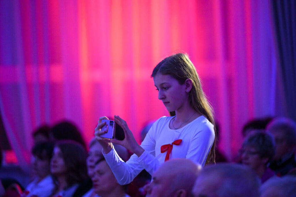 Dziewczyna w białej bluzce z czerwoną kokardą robi zdjęcie telefonem. Tło różowe i fioletowe. W tle siedząca publiczność.