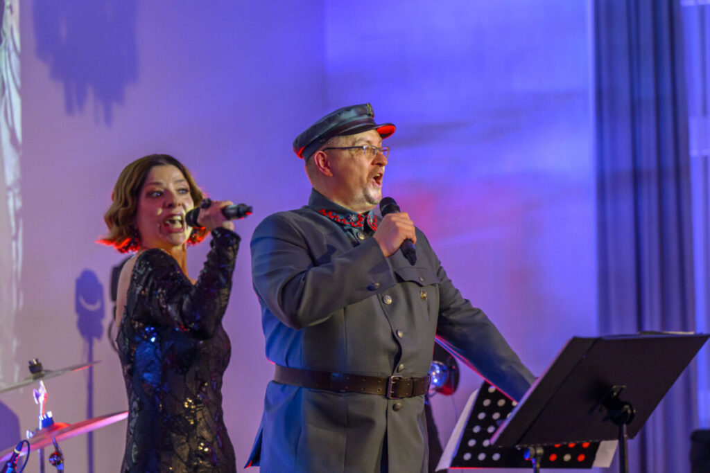 Na scenie śpiewają kobieta i mężczyzna w wojskowym mundurze. Tło jest w kolorze niebiesko-fioletowym. Widać stojak z nutami.