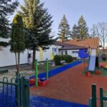 GgC_Tkkh Plac zabaw z niebieską ścieżką i kolorową zjeżdżalnią. W tle białe budynki z czerwoną dachówką. Otoczone zielonymi drzewami.