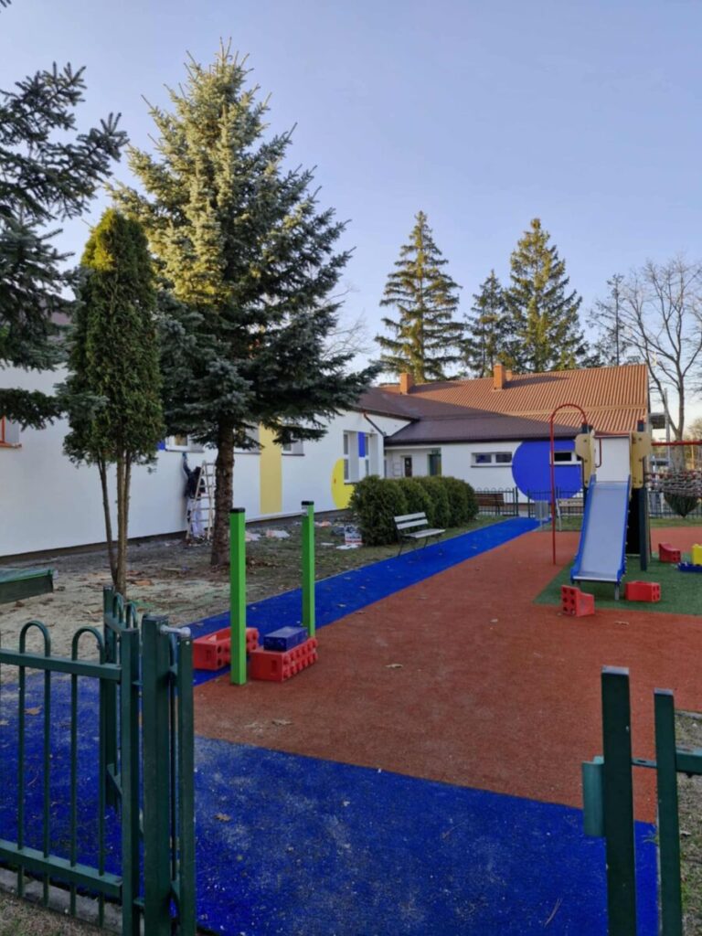 Plac zabaw z niebieską ścieżką i kolorową zjeżdżalnią. W tle białe budynki z czerwoną dachówką. Otoczone zielonymi drzewami.