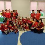 Grupa dzieci i dorosłych ubrana na czerwono na tle jasnych okien. Niebieski dywan na podłodze. Wnętrze sali z jasnymi ścianami.