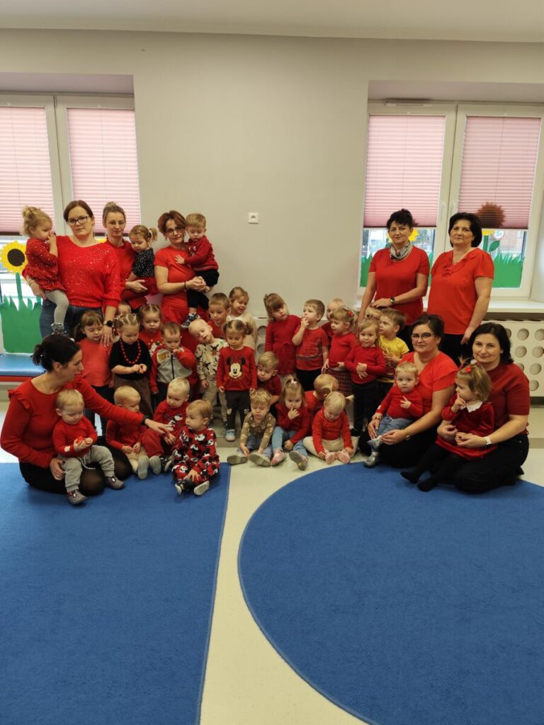 Grupa dzieci i dorosłych ubrana na czerwono na tle jasnych okien. Niebieski dywan na podłodze. Wnętrze sali z jasnymi ścianami.