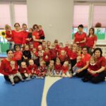 Grupa dzieci i opiekunów w czerwonych ubraniach w sali. W tle szare ściany i okna z różowymi żaluzjami. Na podłodze niebieski dywan.