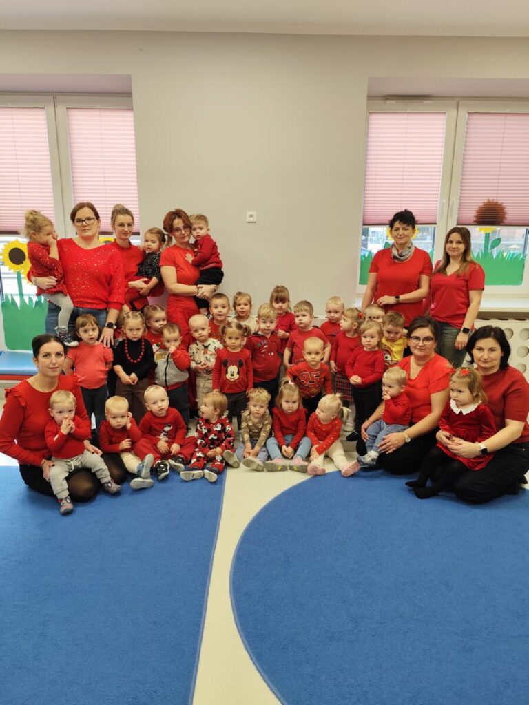 Grupa dzieci i opiekunów w czerwonych ubraniach w sali. W tle szare ściany i okna z różowymi żaluzjami. Na podłodze niebieski dywan.