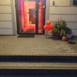 Wejście do budynku z czerwonymi balonami i dekoracjami jesiennymi obok drzwi. Wnętrze podkreślone ciepłym oświetleniem. Na ziemi wyłożony szary chodnik.