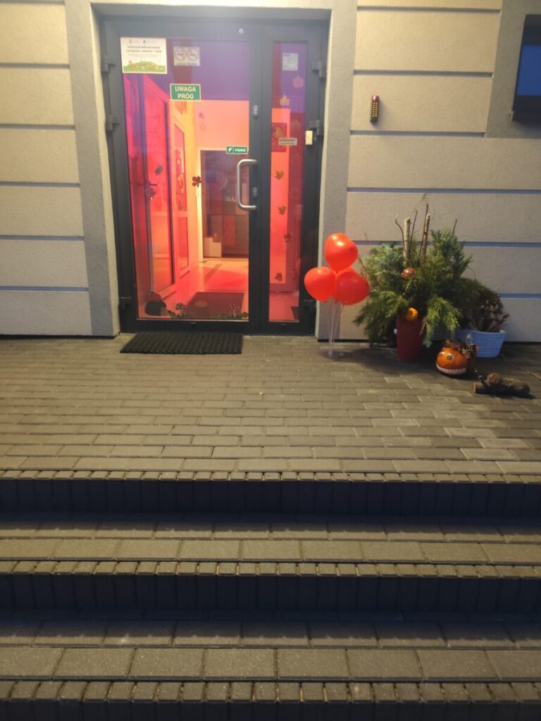Wejście do budynku z czerwonymi balonami i dekoracjami jesiennymi obok drzwi. Wnętrze podkreślone ciepłym oświetleniem. Na ziemi wyłożony szary chodnik.