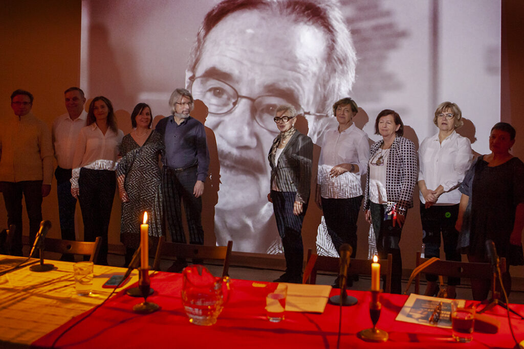 Grupa osób w jasnych strojach stoi przed czarno-białą projekcją twarzy starszego mężczyzny. Na pierwszym planie stół z czerwonym obrusem i świecami. Ciepłe oświetlenie tworzy przytulną atmosferę.