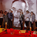 Grupa osób w jasnych strojach stoi przed czarno-białą projekcją twarzy starszego mężczyzny. Na pierwszym planie stół z czerwonym obrusem i świecami. Ciepłe oświetlenie tworzy przytulną atmosferę.