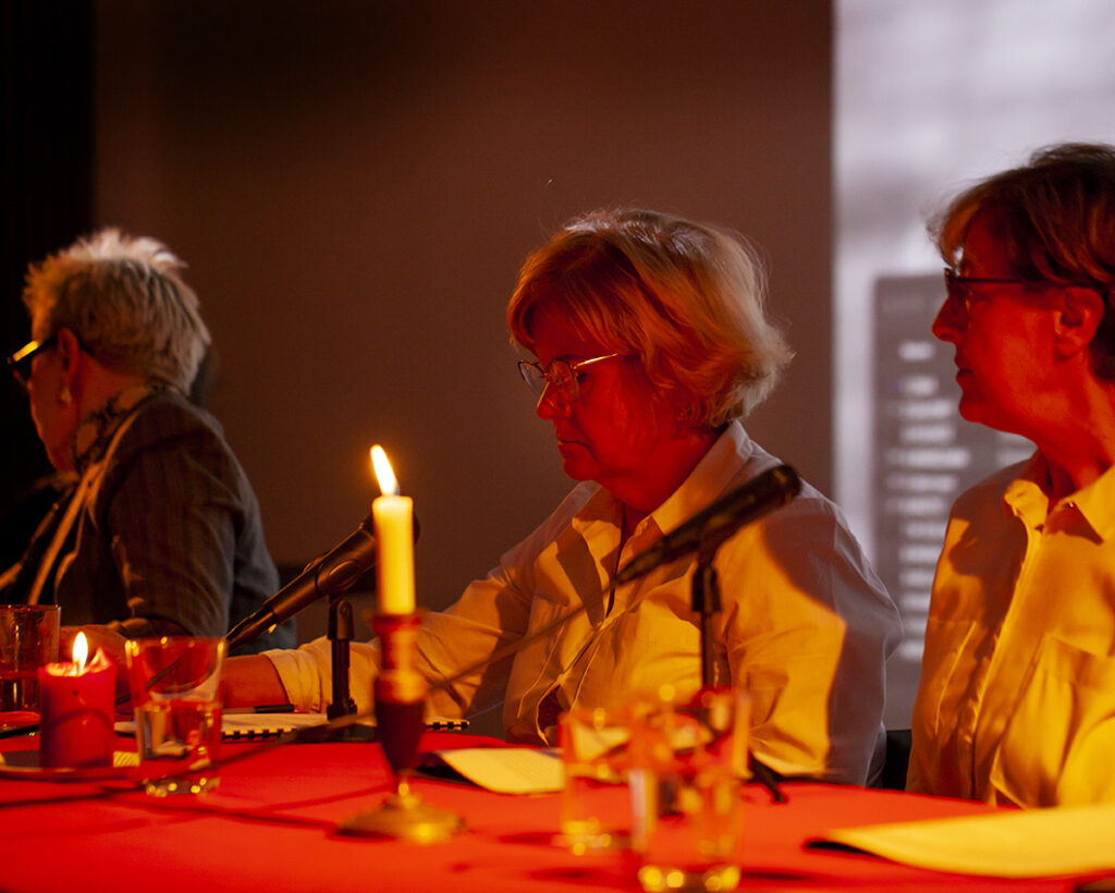 Trzy osoby siedzą przy stole w ciemnym pomieszczeniu. Stół pokryty jest czerwonym obrusem i oświetlony świecami. W tle ekran z rozmazanym obrazem.