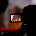 Kamera nagrywa dwie osoby w jasnym świetle. W tle ciepłe, pomarańczowe kolory. Dwóch mówców przy mikrofonie w obiektywie.