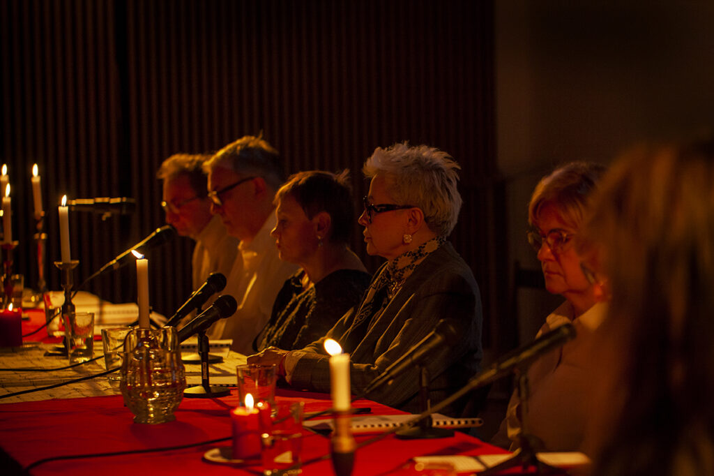 Zdjęcie pokazuje grupę osób przy stole z czerwonym obrusem, oświetlonych świecami. Stoły są ozdobione świecami w ciemnym pomieszczeniu. Osoby siedzą przy mikrofonach, co wskazuje na dyskusję lub spotkanie.