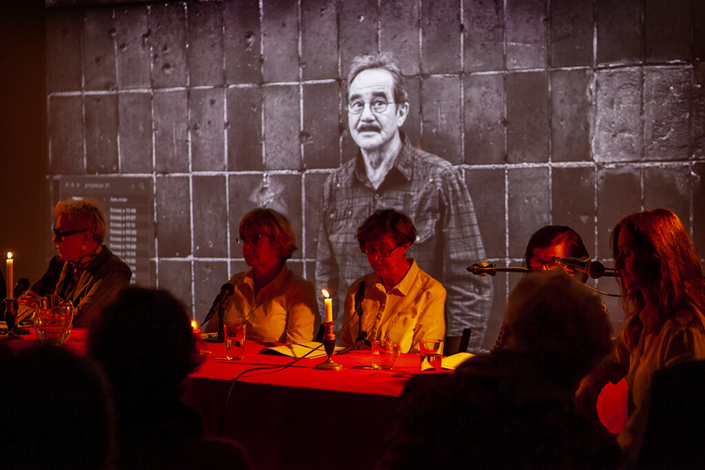 Grupa osób siedzi przy stole w ciemnym pomieszczeniu, oświetlonym świecami. W tle widoczne jest czarno-białe zdjęcie mężczyzny na tle ściany. Przewaga ciemnych, ciepłych kolorów.