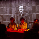 Grupa osób siedzi przy stole w ciemnym pomieszczeniu, oświetlonym świecami. W tle widoczne jest czarno-białe zdjęcie mężczyzny na tle ściany. Przewaga ciemnych, ciepłych kolorów.