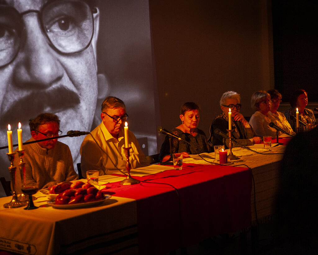 Na zdjęciu widać grupę osób siedzących przy stole, na którym palą się świece. Tło stanowi wielki czarno-biały portret. Atmosfera jest ciepła, z dominacją ciepłych odcieni.