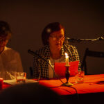 Dwie kobiety siedzą przy stole z czerwonym obrusem, każda z mikrofonem. Na stole paląca się świeca i szklanki. Tło jest ciemne.