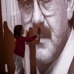 Małe dziecko dotyka czarno-białego zdjęcia twarzy mężczyzny na ścianie. Obraz jest duży, a postać nosi okulary. Dziecko jest w czerwonej bluzce i szarych spodniach.