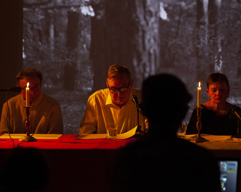 Trzy osoby siedzą przy stole, oświetlone świecami. W tle czarno-białe zdjęcie lasu. Ciepłe, przytłumione światło dominuje obraz.