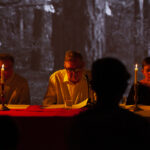 Trzy osoby siedzą przy stole, oświetlone świecami. W tle czarno-białe zdjęcie lasu. Ciepłe, przytłumione światło dominuje obraz.