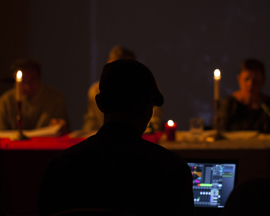 Na ciemnym tle widać osobę przy komputerze, przed nią palą się świece. W tle rozmyte postacie siedzą przy stole. Ciepłe, żółte światło dominuje na obrazie.