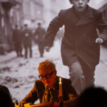 Osoba siedzi przy stole z mikrofonem i świecą. W tle czarno-białe zdjęcie biegającego chłopca na ulicy. Scena jest oświetlona ciepłym światłem.