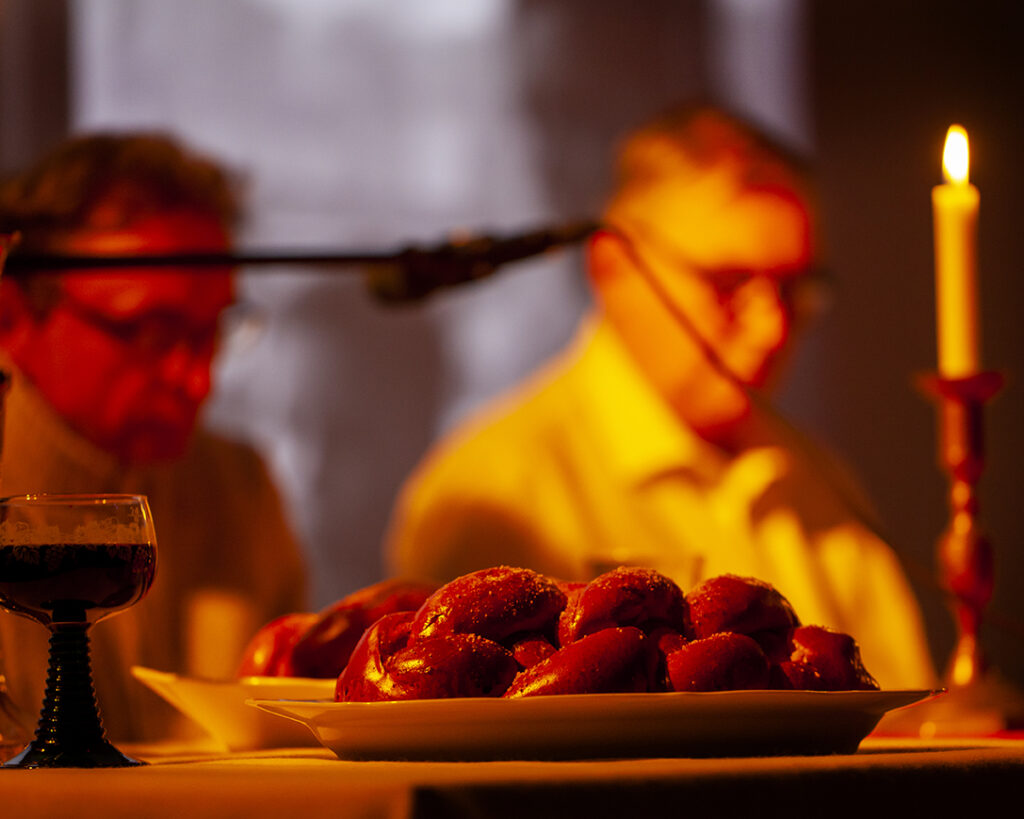 Na stole leżą świeże bułki w pomarańczowym świetle świecy. W tle dwie osoby rozmawiają przy stole. Obok stoi kielich z ciemnym napojem.