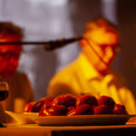 Na stole leżą świeże bułki w pomarańczowym świetle świecy. W tle dwie osoby rozmawiają przy stole. Obok stoi kielich z ciemnym napojem.