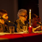 Na zdjęciu kilka osób siedzi przy stole oświetlonym świecą. Mają mikrofony i czytają dokumenty. Tło jest ciemne.