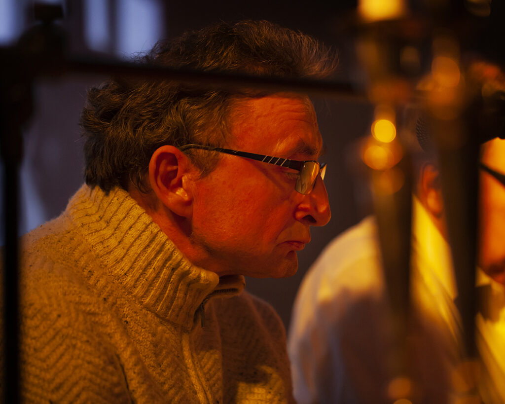 Starszy mężczyzna w okularach, ubrany w beżowy sweter, siedzi przy złotym oświetleniu. Tło jest przyciemnione, co nadaje ciepły nastrój. Obok widoczne są rozmyte sylwetki.