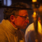 Starszy mężczyzna w okularach, ubrany w beżowy sweter, siedzi przy złotym oświetleniu. Tło jest przyciemnione, co nadaje ciepły nastrój. Obok widoczne są rozmyte sylwetki.