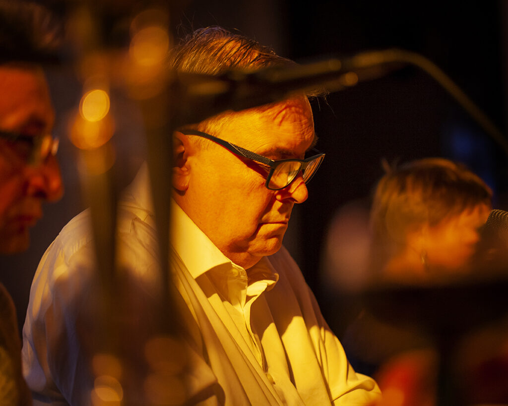 Nie rozpoznaję osób na zdjęciu. Obraz przedstawia mężczyznę w okularach, w białej koszuli, w ciepłym, żółtym świetle. W tle znajduje się niewyraźnie zarysowana postać, a atmosfera jest przytulna i intymna.