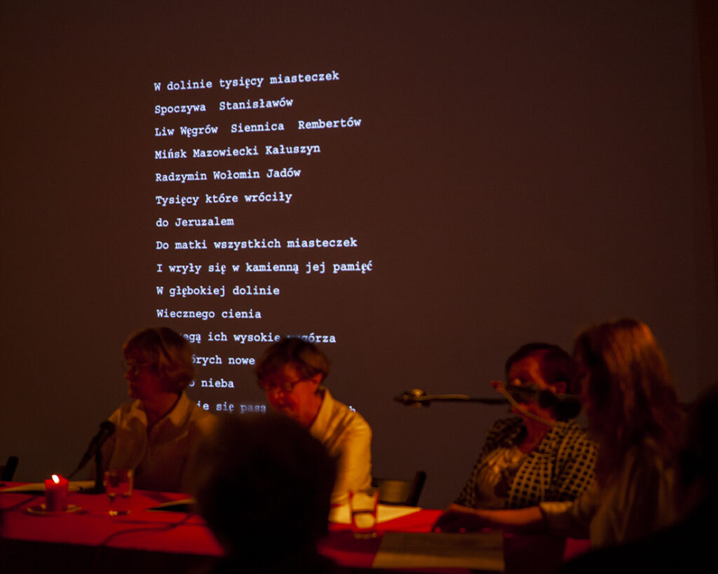 W ciemnym pomieszczeniu, na ekranie wyświetlony jest biały tekst. Przy czerwonym stole siedzą cztery osoby przy mikrofonach. Atmosfera jest poważna.