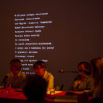 W ciemnym pomieszczeniu, na ekranie wyświetlony jest biały tekst. Przy czerwonym stole siedzą cztery osoby przy mikrofonach. Atmosfera jest poważna.