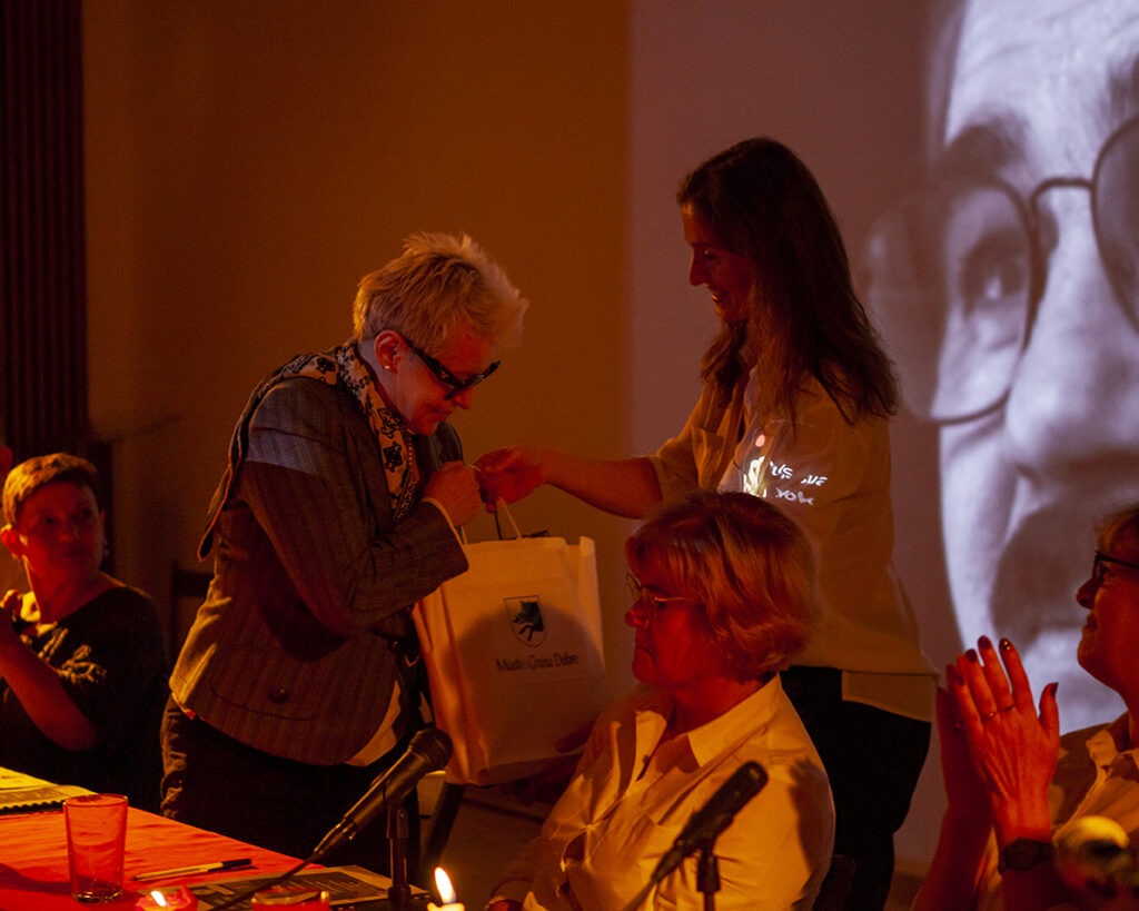 Na zdjęciu widać starszą kobietę otrzymującą torbę podczas wydarzenia w ciepłym oświetleniu. W tle jest duża projekcja twarzy. Kobiety siedzą przy stole z mikrofonem, a atmosfera jest przyjazna.