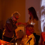 Na zdjęciu widać starszą kobietę otrzymującą torbę podczas wydarzenia w ciepłym oświetleniu. W tle jest duża projekcja twarzy. Kobiety siedzą przy stole z mikrofonem, a atmosfera jest przyjazna.