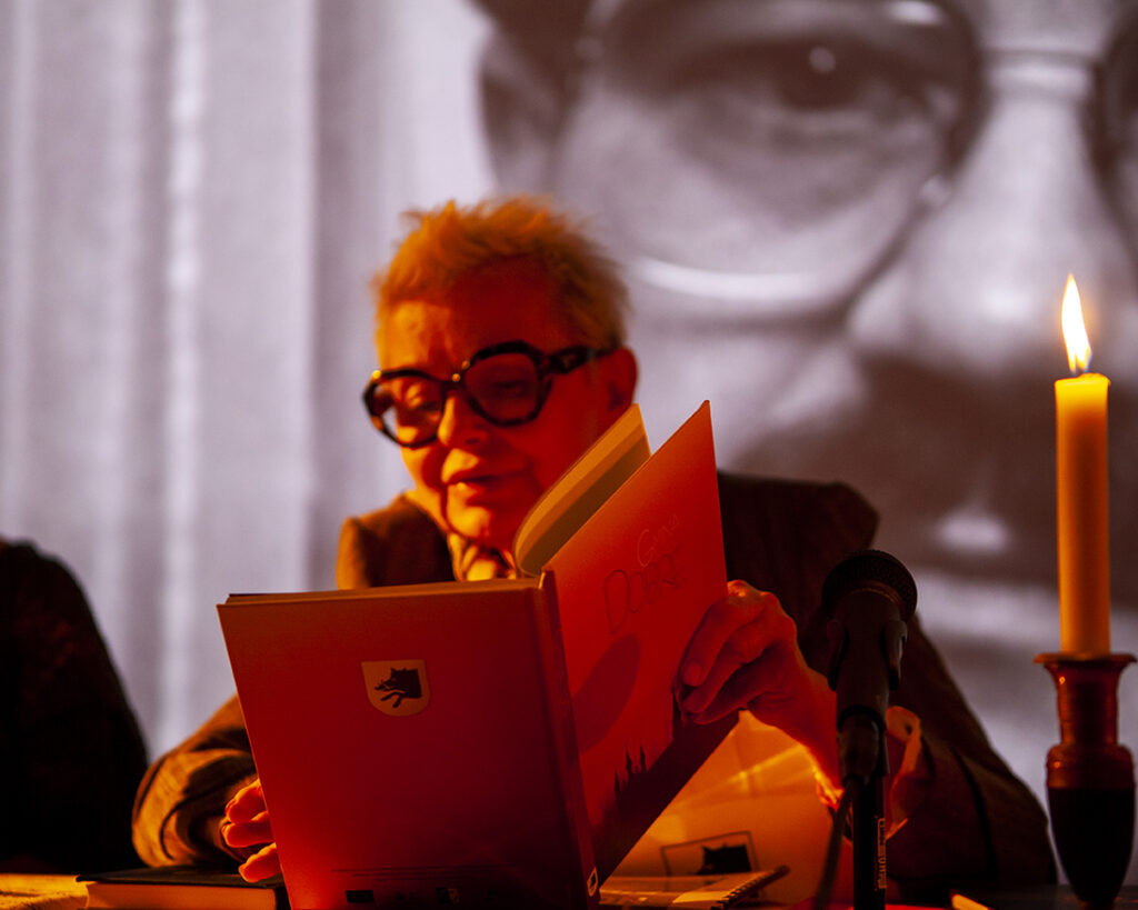 Osoba w okularach czyta książkę przy świecy, tło jest czarno-białe z dużym portretem. Przeważają odcienie pomarańczowe.