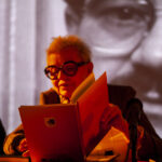 Osoba w okularach czyta książkę przy świecy, tło jest czarno-białe z dużym portretem. Przeważają odcienie pomarańczowe.