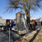 Na zdjęciu ludzie pracują przy małej kapliczce obok drzewa. Kapliczka jest szara, z drabiną opartą o nią. W tle jasnoniebieskie niebo i samochód dostawczy.