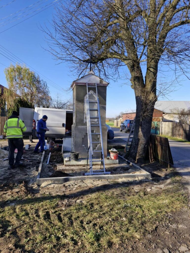 Na zdjęciu widać szary pomnik z drabiną na tle błękitnego nieba i dużego drzewa. Obok pracują dwaj mężczyźni w jaskrawych kurtkach. W tle stoi biały samochód dostawczy.