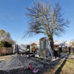 Plac budowy przy kapliczce z drabiną, obok duże drzewo bez liści. Pracownik w pomarańczowej kamizelce stoi przy białym vanem. Tło stanowi niebieskie niebo.