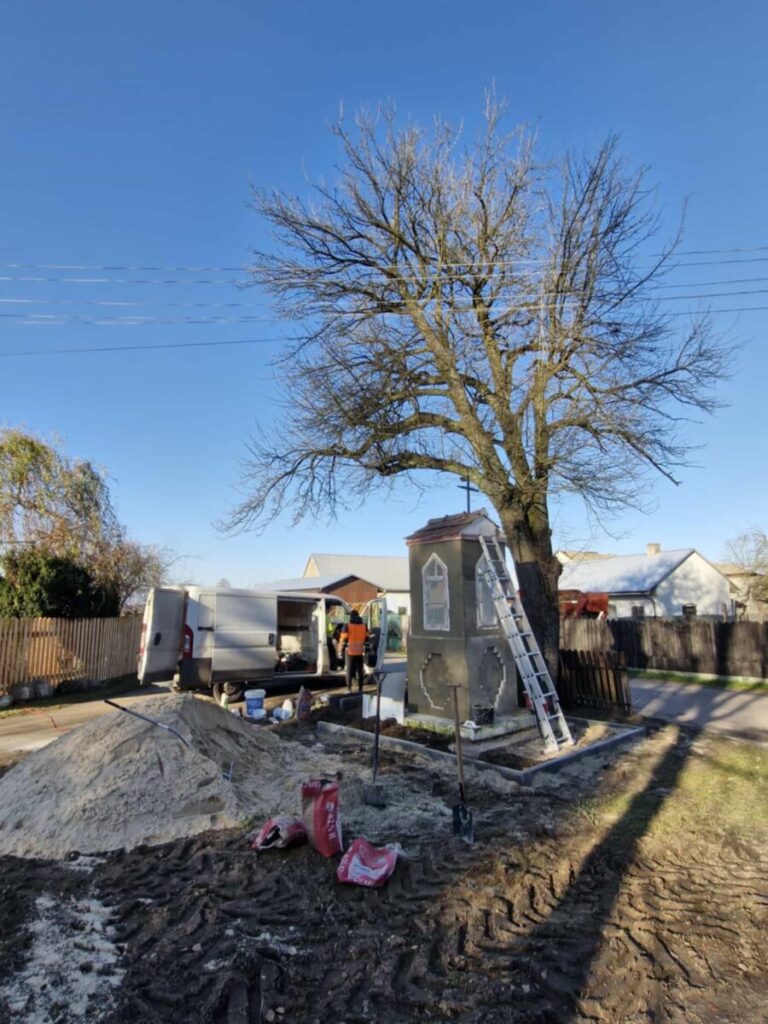 Plac budowy przy kapliczce z drabiną, obok duże drzewo bez liści. Pracownik w pomarańczowej kamizelce stoi przy białym vanem. Tło stanowi niebieskie niebo.