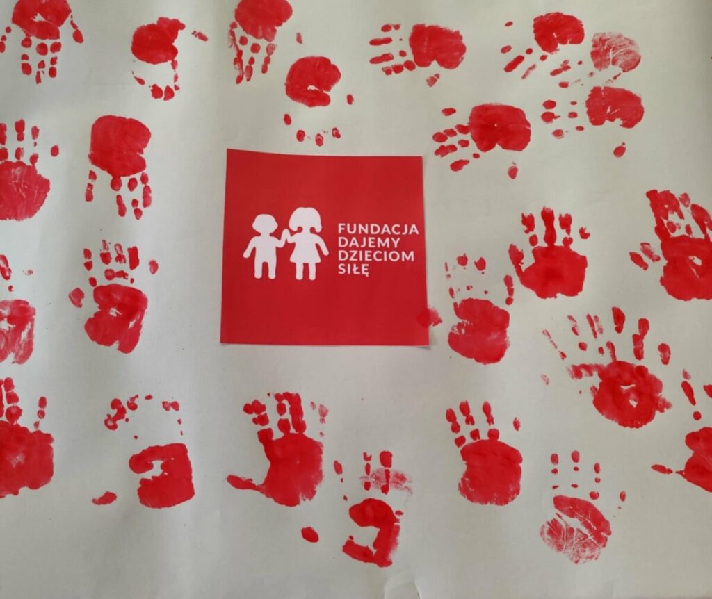 Czerwone odciski dłoni na białym tle tworzą wzór wokół logo fundacji z dziećmi. Centralnie plakietka z napisem. Dominują kolory czerwony i biały.