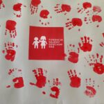 Czerwone odciski dłoni na białym tle tworzą wzór wokół logo fundacji z dziećmi. Centralnie plakietka z napisem. Dominują kolory czerwony i biały.