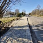 Chodnik z kostki brukowej biegnie wzdłuż drogi asfaltowej, po lewej stronie drzewa i trawnik. Słoneczny dzień, jasnoniebieskie niebo. W tle widoczny zaparkowany samochód i domy.