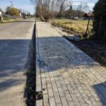 Chodnik z jasnych płytek biegnie wzdłuż asfaltowej drogi. Po prawej są drzewa i trawniki, w tle widać niebo i chmury.