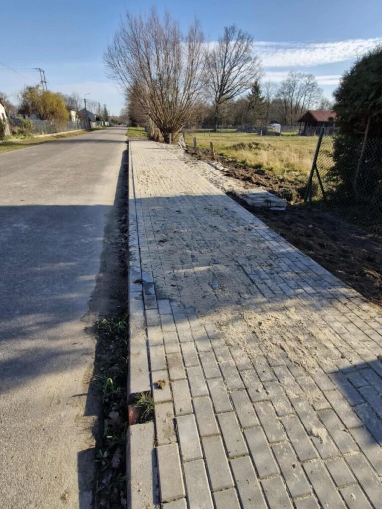 Chodnik z jasnych płytek biegnie wzdłuż asfaltowej drogi. Po prawej są drzewa i trawniki, w tle widać niebo i chmury.